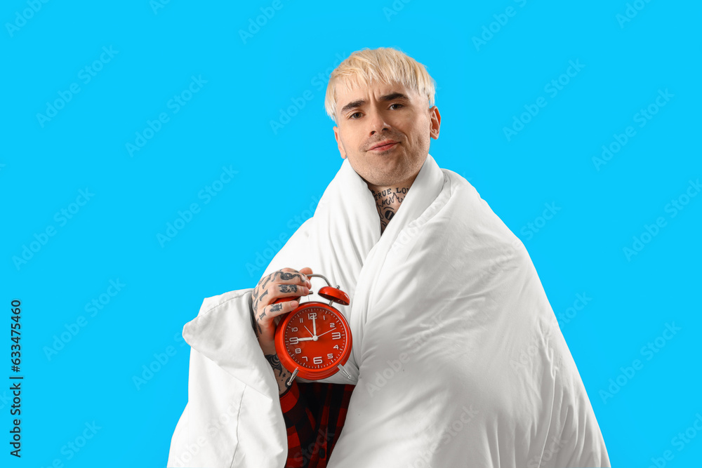 Young tattooed man with blanket and alarm clock on blue background