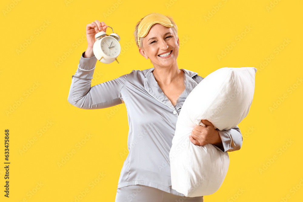 Mature woman in pajamas with pillow and alarm clock on yellow background