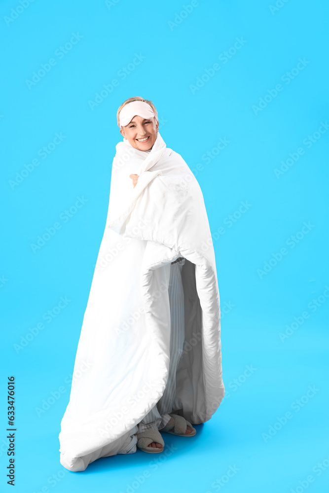 Mature woman with sleeping mask and blanket on blue background