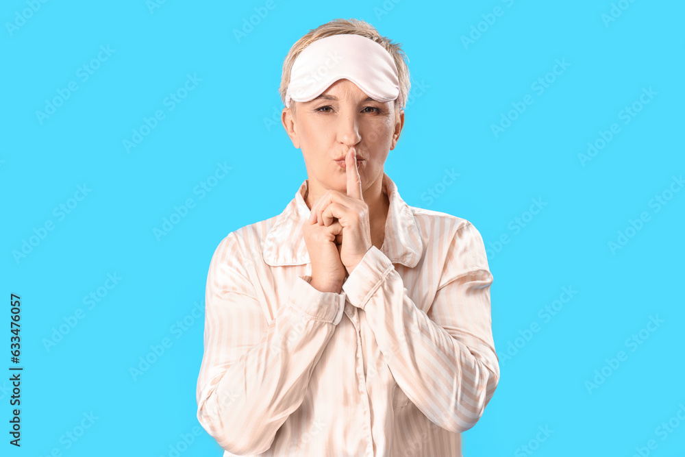 Mature woman in pajamas with sleeping mask showing silence gesture on blue background