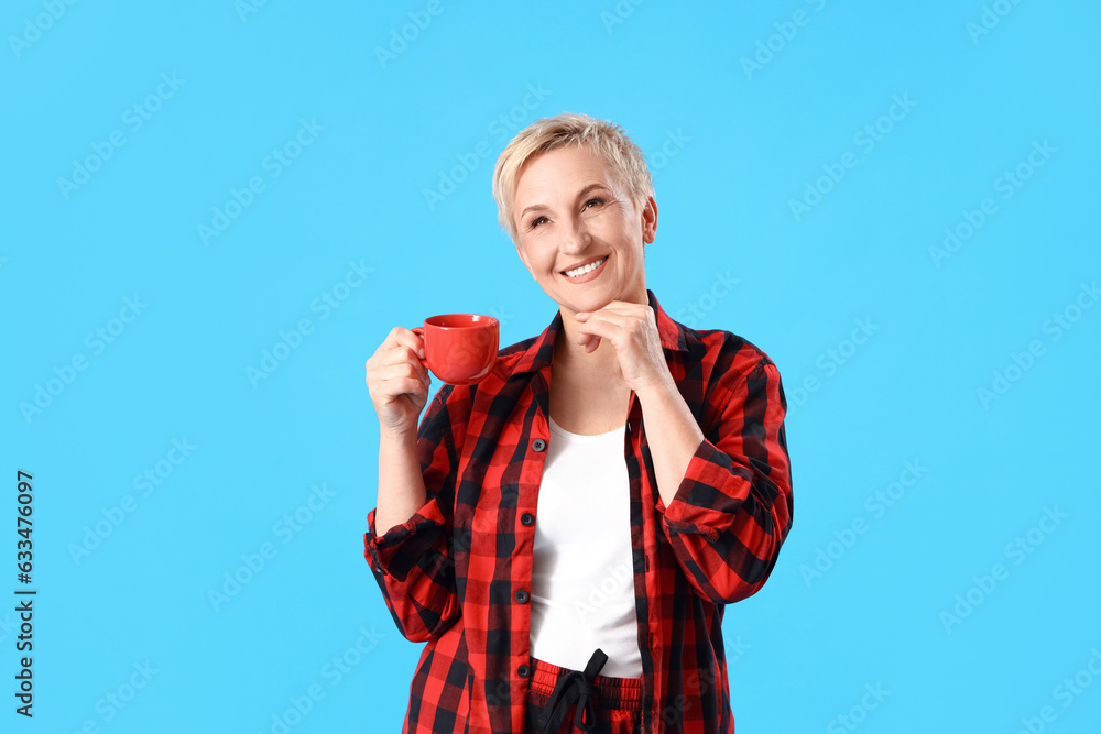 Mature woman in pajamas with cup of coffee on blue background