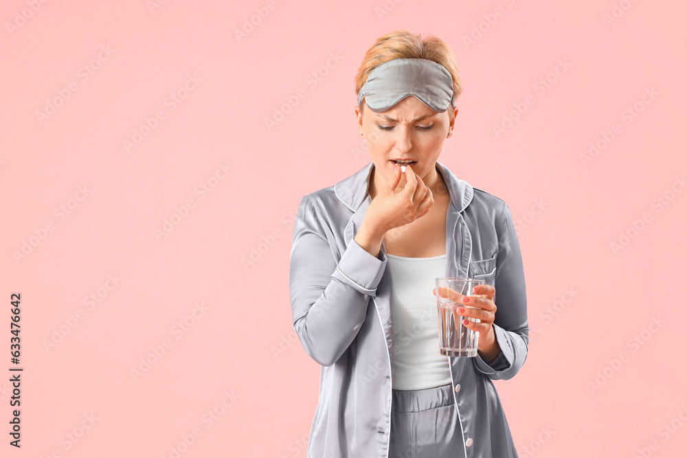 Young woman in pajamas with glass of water taking pill on pink background