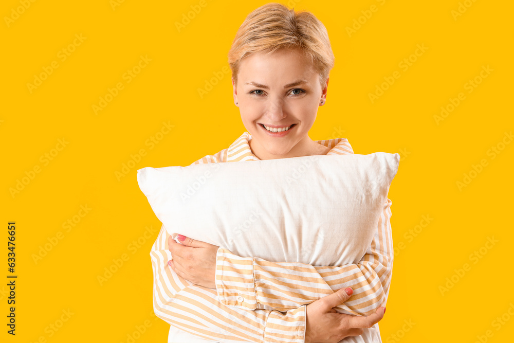 Young woman in pajamas with pillow on yellow background