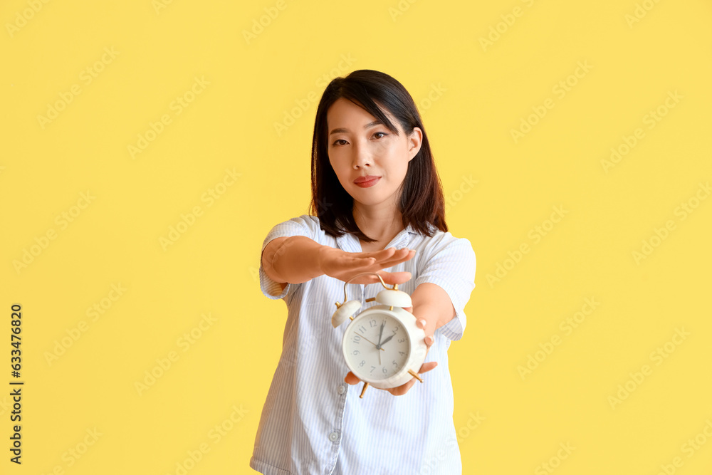 Beautiful Asian woman in pajamas with alarm clock on yellow background