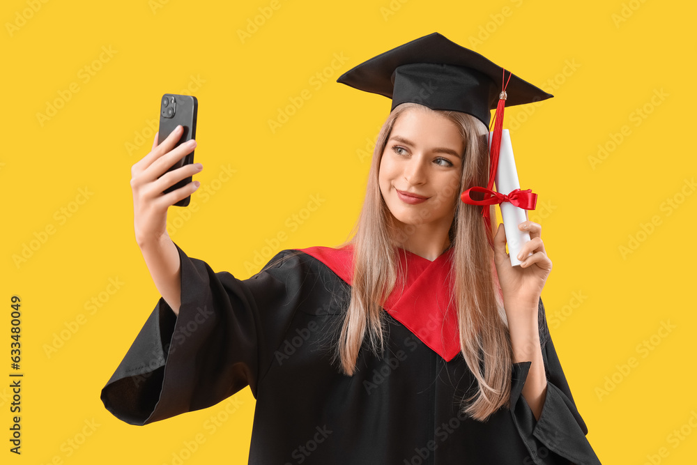 Female graduate student with diploma taking selfie on yellow background