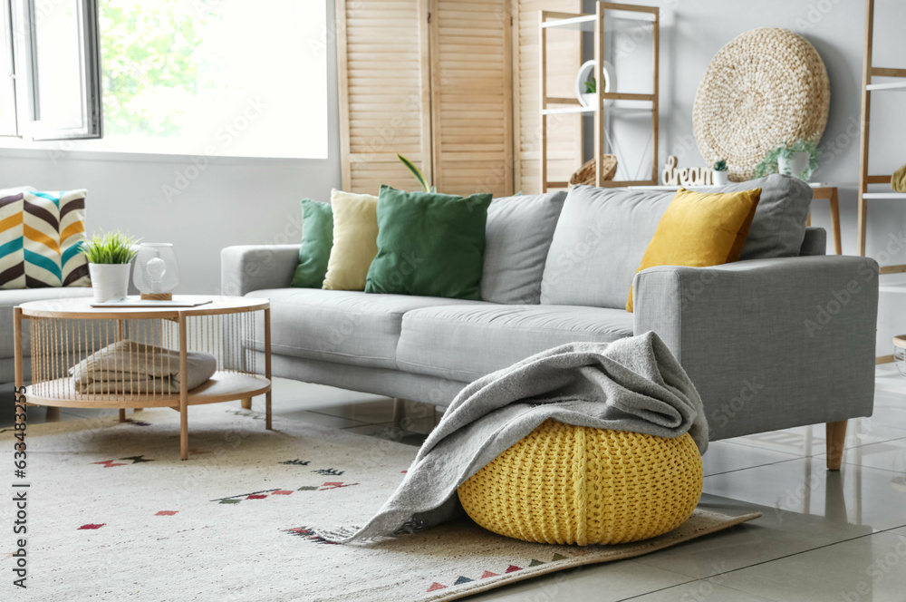 Cozy grey sofa, coffee table and pouf with soft blanket in interior of living room