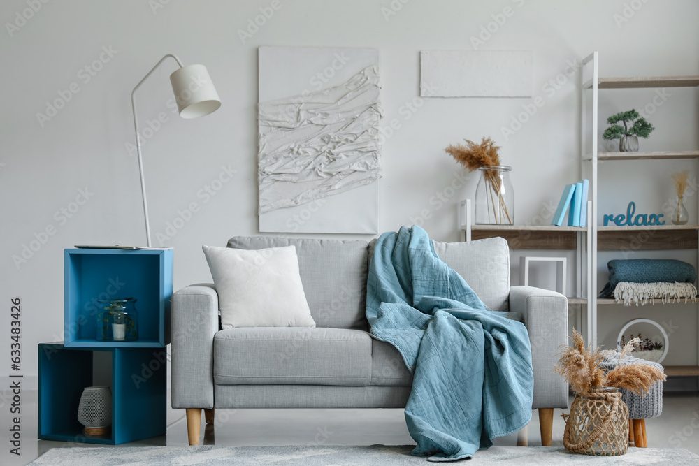 Cozy grey sofa with soft blanket, cushion and stylish shelves in interior of light living room