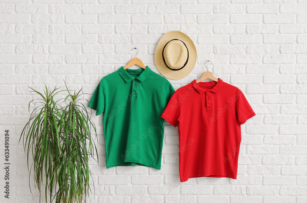 Stylish t-shirts and hat hanging on white brick wall