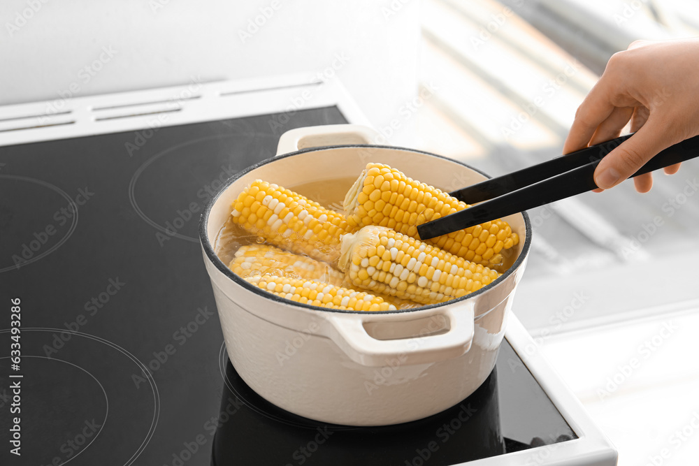 Woman boiling corn cobs in cooking pot on electric stove in kitchen