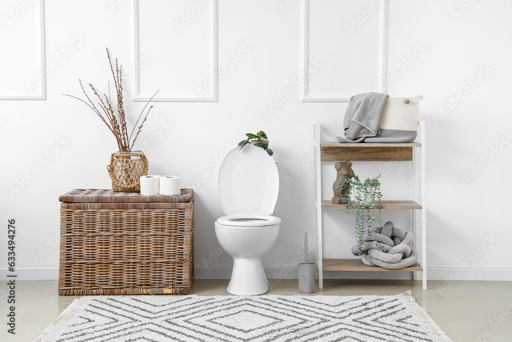 Interior of light restroom with ceramic toilet bowl, shelving unit and wicker basket