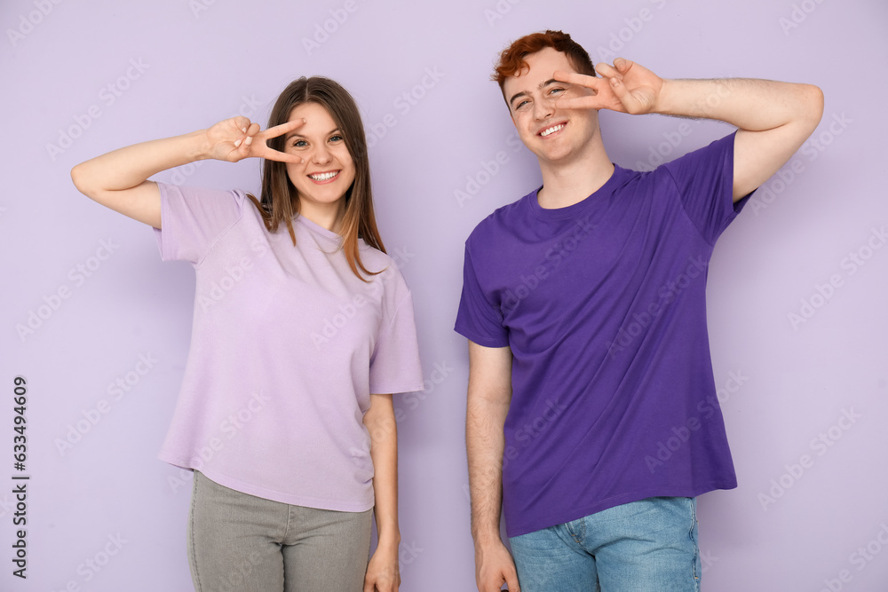 Young couple in t-shirts showing victory gesture on lilac background