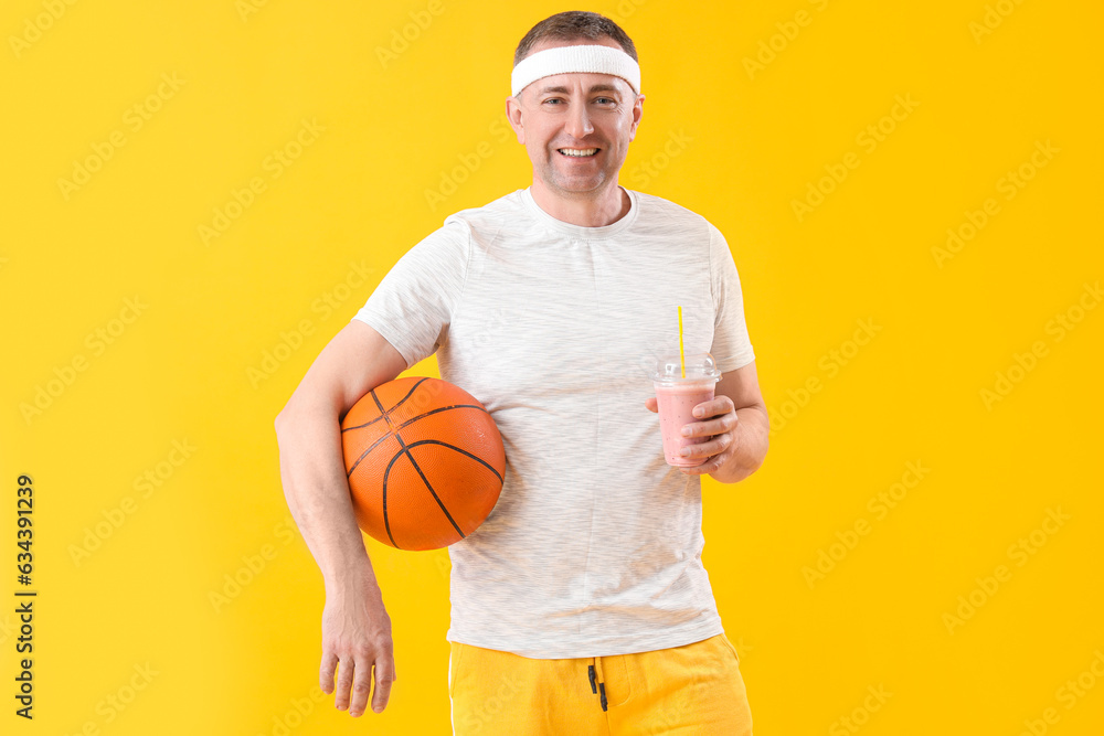 Sporty mature man with glass of fruit smoothie and ball on yellow background