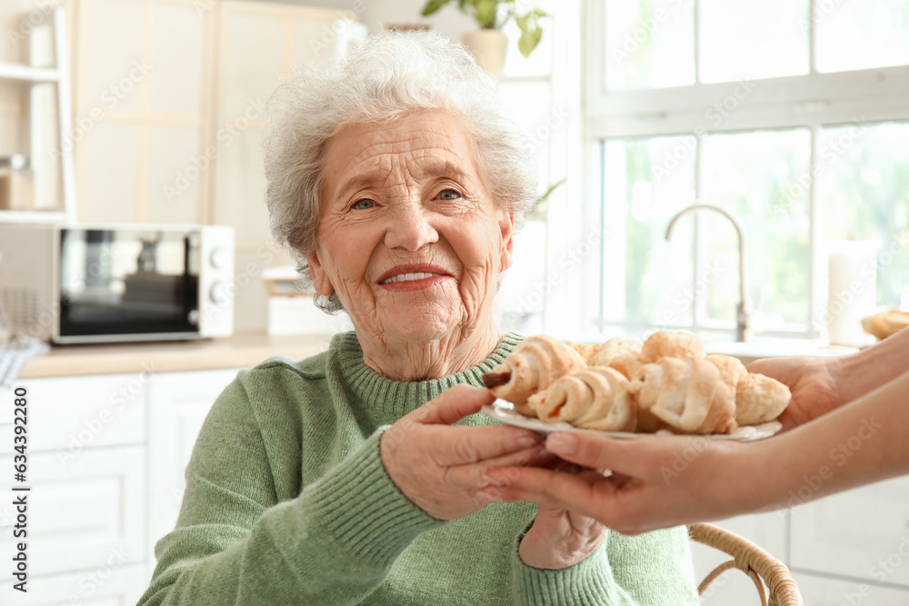 Young woman giving tasty croissants to her grandmother in kitchen, closeup