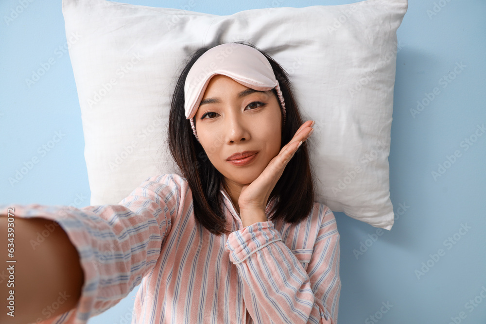 Beautiful Asian woman in pajamas with pillow taking selfie on blue background