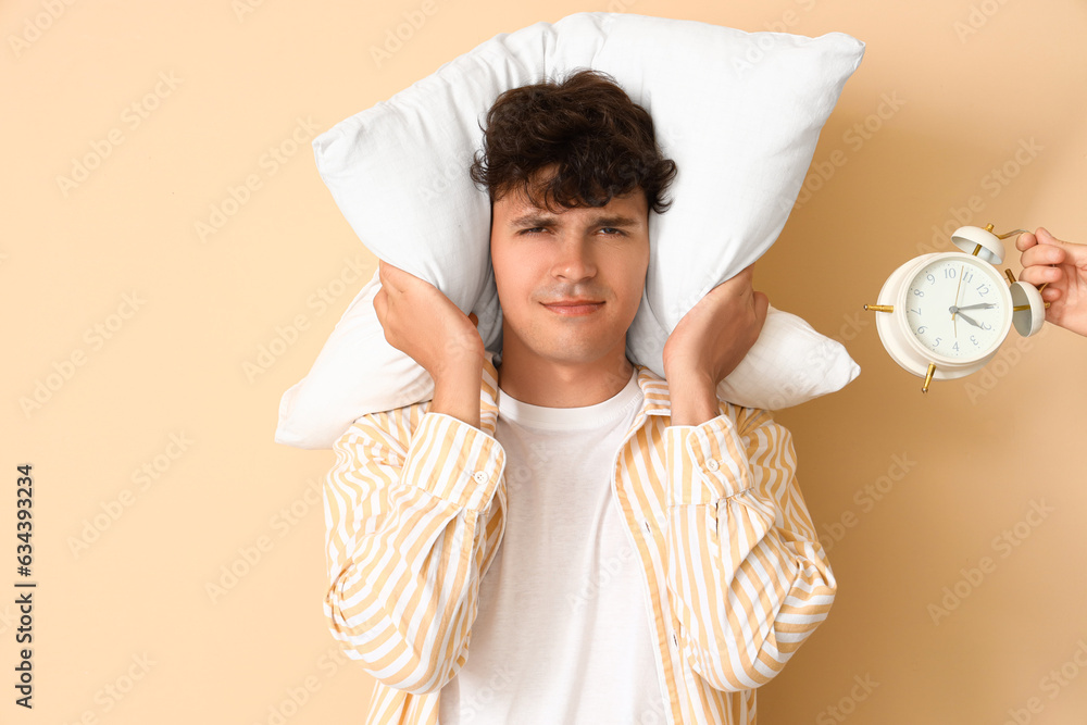 Displeased young man with pillow and ringing alarm clock on beige background