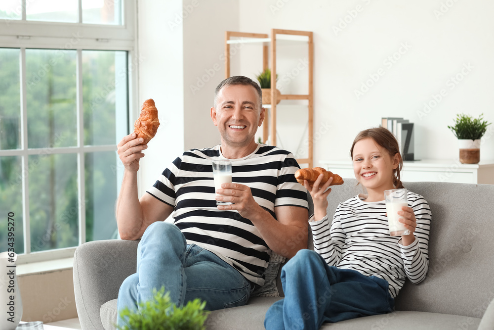 Little girl and her father with croissants drinking milk in living room