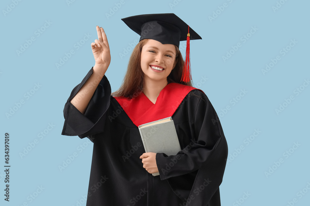 Female graduate student with book showing victory gesture on blue background