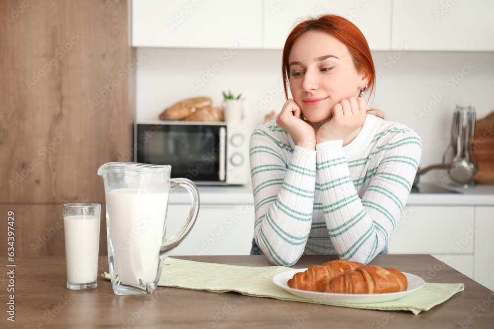 Young redhead woman with glass of milk and croissants in kitchen