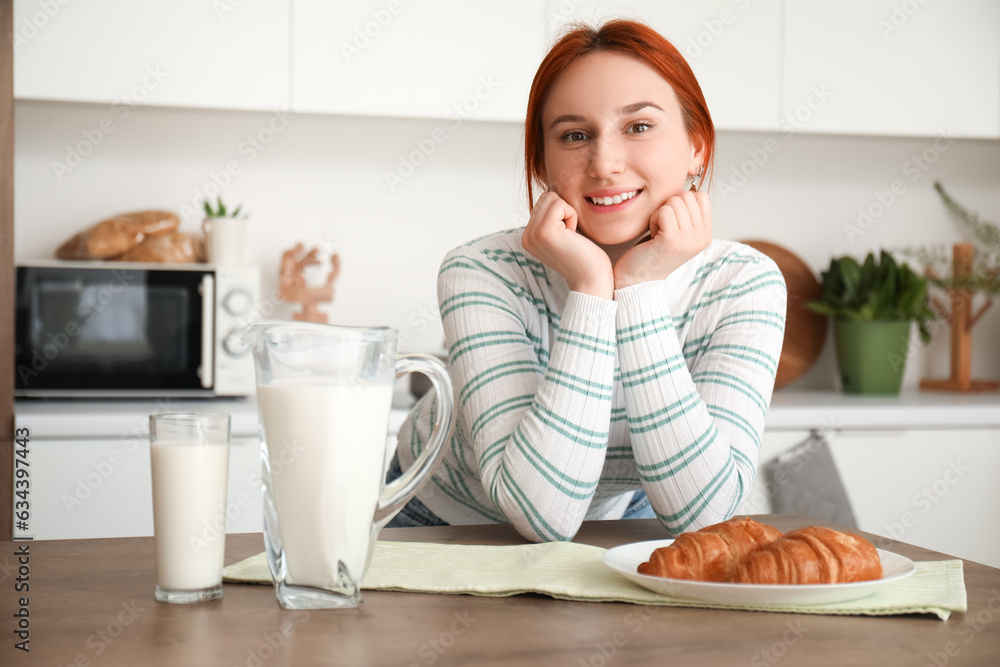 Young redhead woman with glass of milk and croissants in kitchen