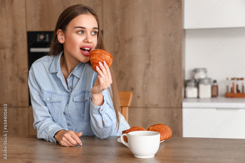 Beautiful young woman with cup of coffee eating croissant in kitchen