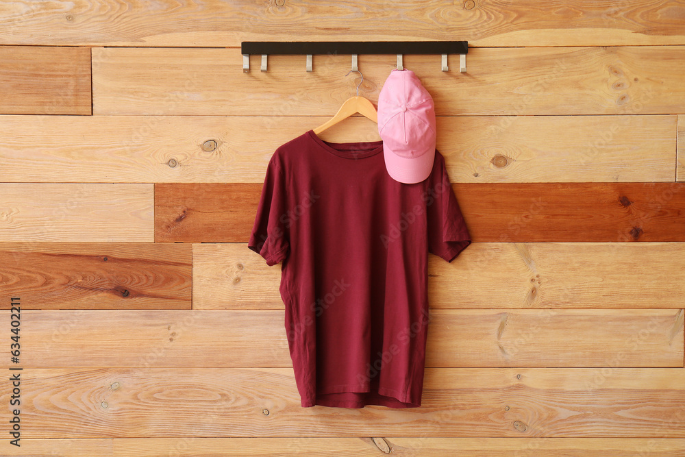 Stylish red t-shirt and cap hanging on wooden background