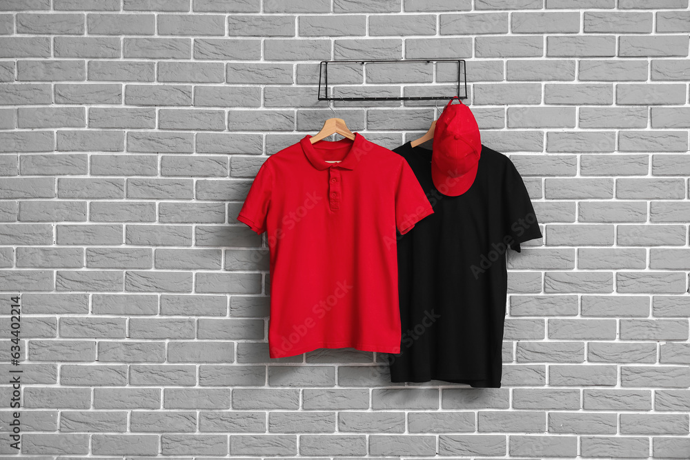 Stylish t-shirts and cap hanging on grey brick wall