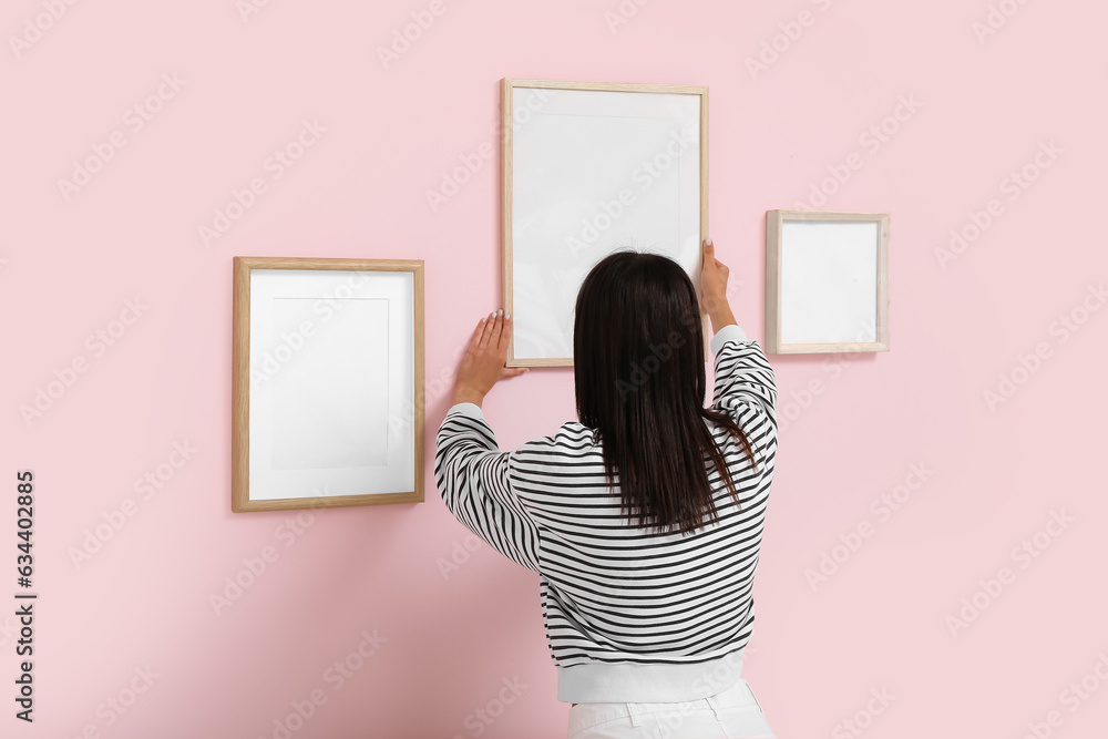 Young woman hanging blank frame on pink wall in living room, back view