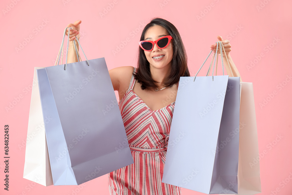 Happy beautiful young Asian woman in sunglasses with shopping bags on pink background