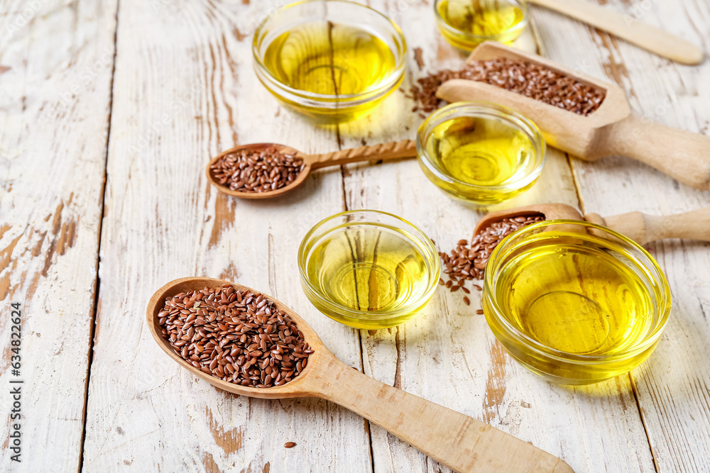 Glass bowls of flax oil and spoons with seeds on white wooden background