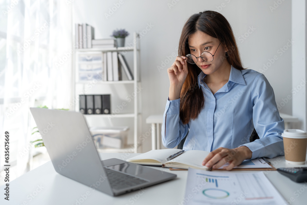 Confident asian businesswoman sitting and taking notes in notebook from laptop computer on financial