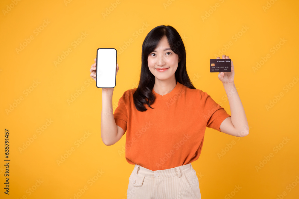 Vibrant Asian woman in her 30s, holding smartphone and credit card on yellow background. Hassle-free