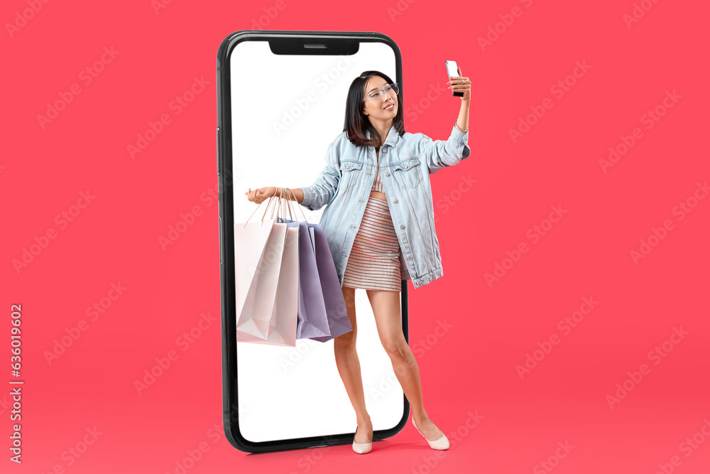 Stylish Asian woman with shopping bags taking selfie near big mobile phone on red background