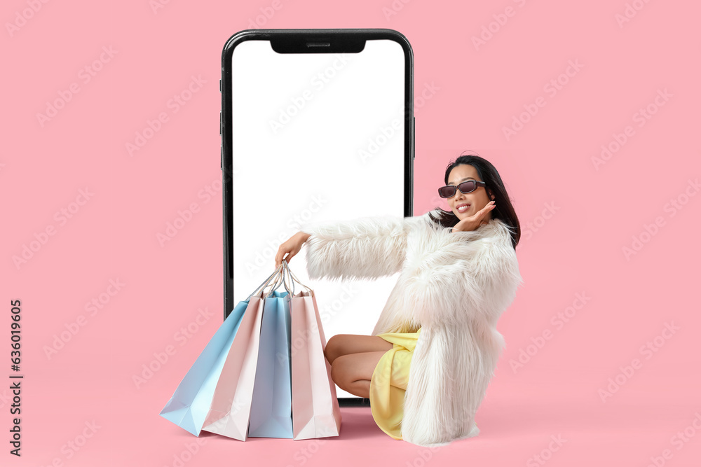 Stylish Asian woman with shopping bags and big mobile phone on pink background