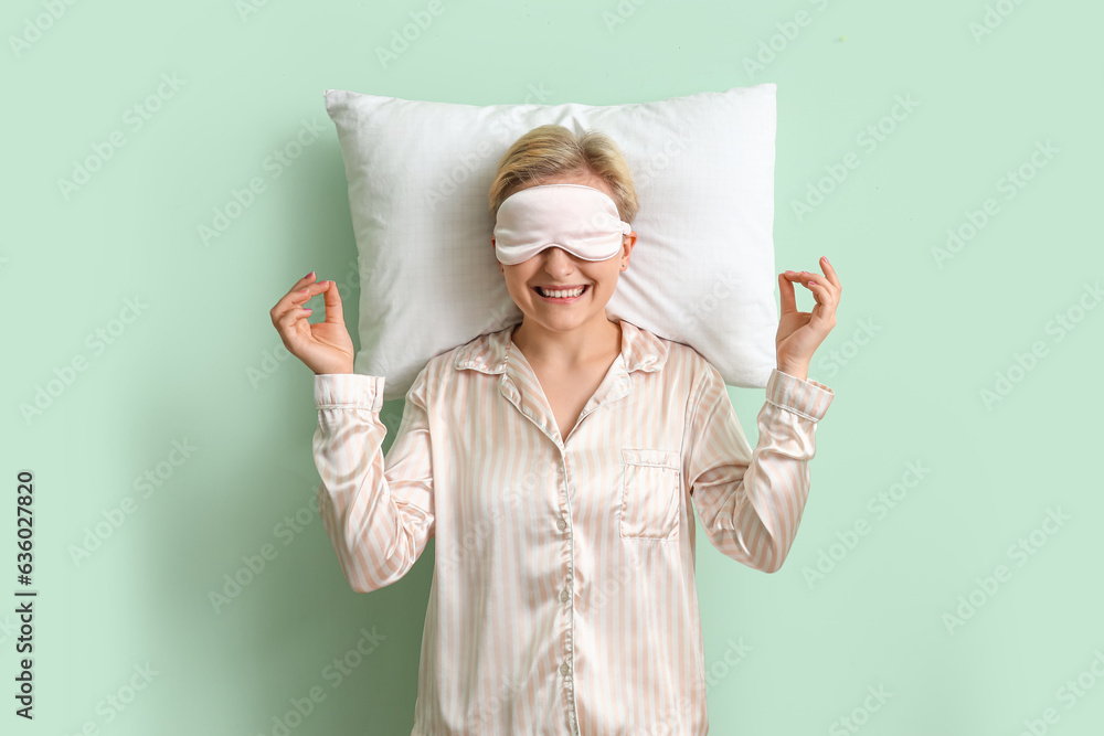 Young woman in pajamas with pillow meditating on green background