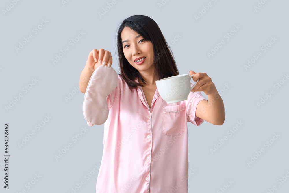 Beautiful Asian woman in pajamas with sleeping mask and coffee cup on grey background