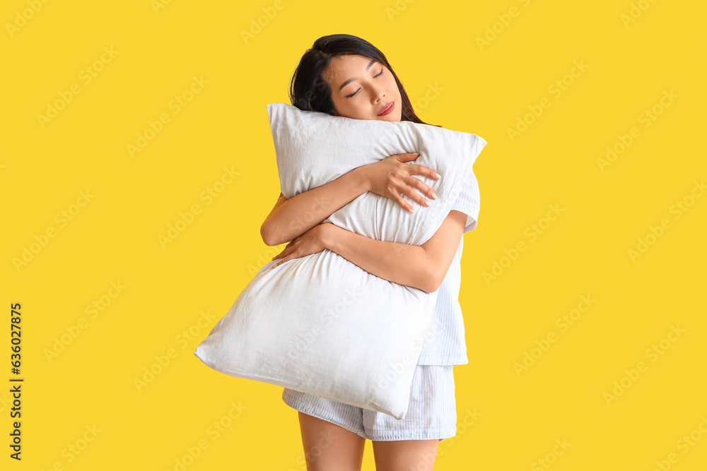 Beautiful Asian woman in pajamas with pillow on yellow background