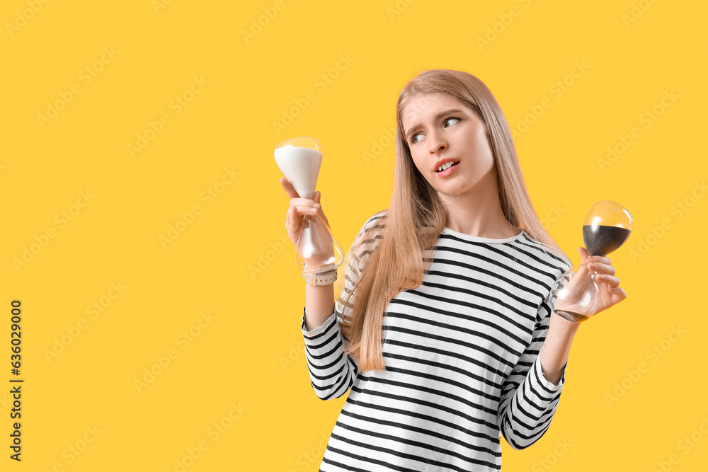 Stressed young woman with hourglass on yellow background. Deadline concept