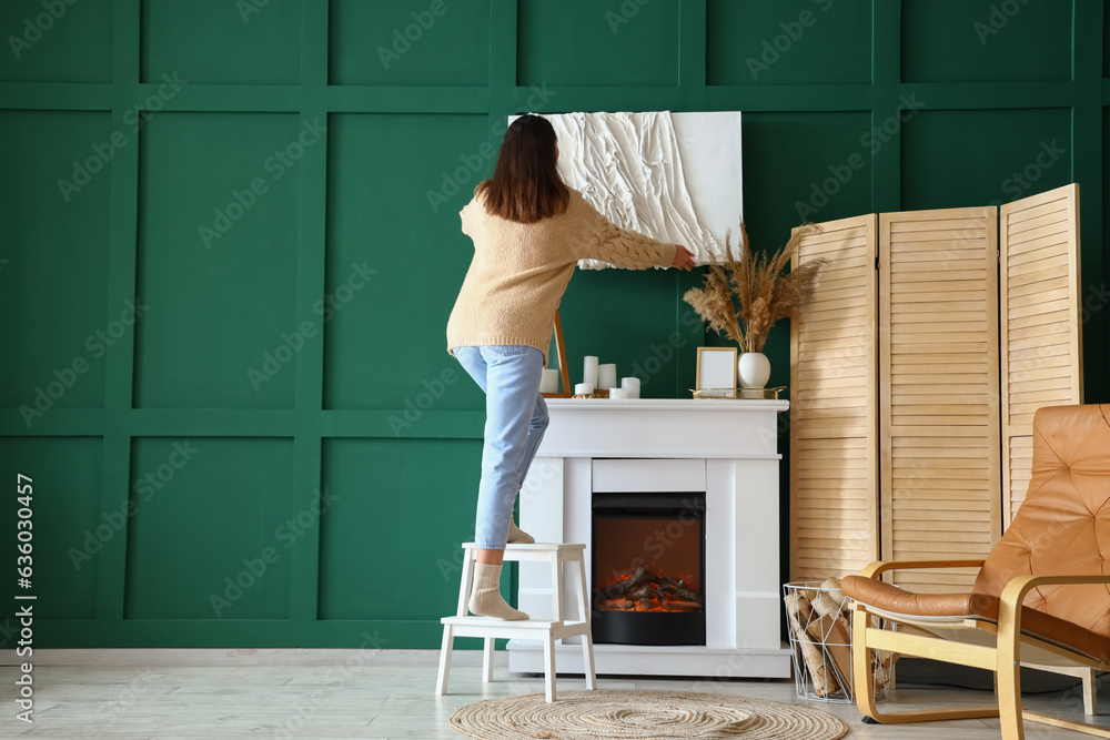 Young woman hanging artwork on green wall above fireplace at home, back view