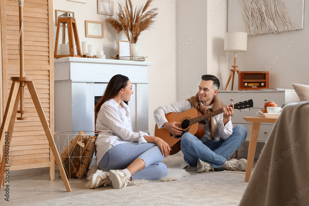 Young woman and her husband playing guitar near fireplace at home