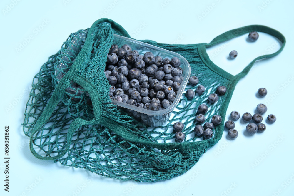 Mesh bag with fresh blueberry in plastic container on blue background, closeup