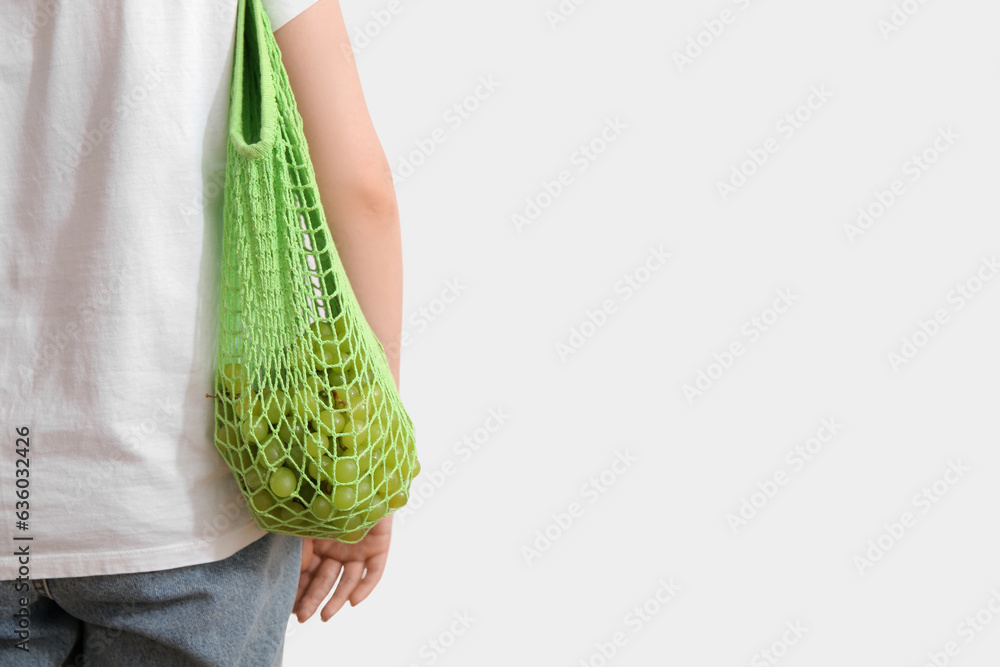 Woman with fresh grapes in mesh bag on light background, back view