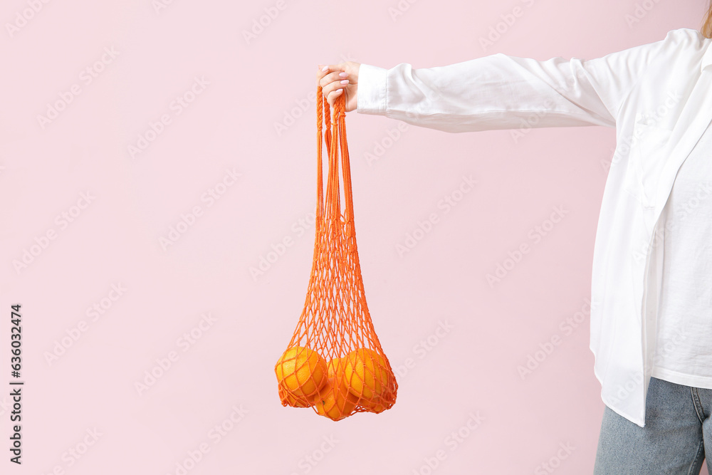 Female hand holding mesh bag with fresh oranges on beige background