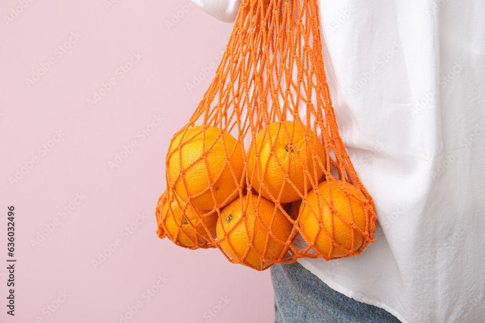 Woman with fresh oranges in mesh bag on beige background, closeup