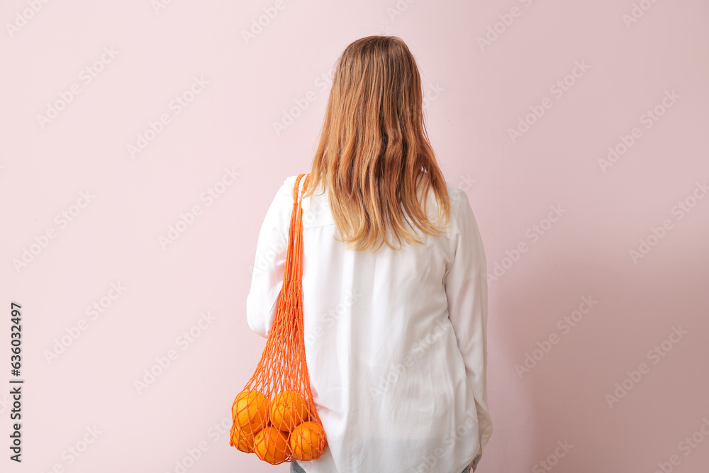 Woman with fresh oranges in mesh bag on beige background, back view