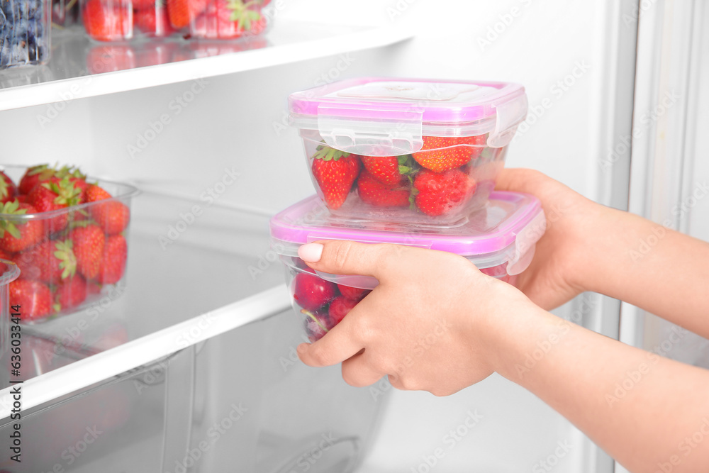 Female hands and containers with fresh strawberry near refrigerator, closeup