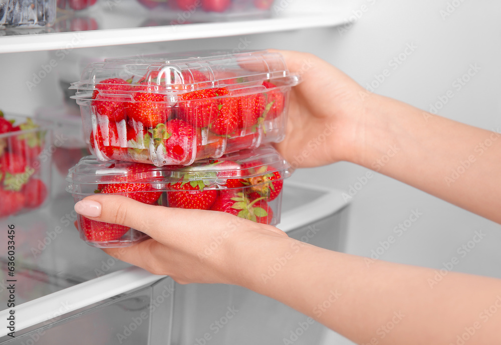 Woman holding plastic containers with fresh strawberry near refrigerator, closeup