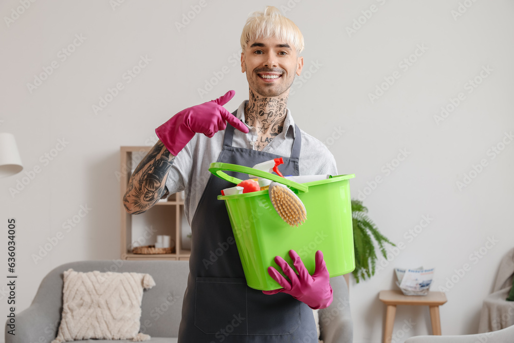 Young tattooed man pointing at bucket of cleaning supplies in room