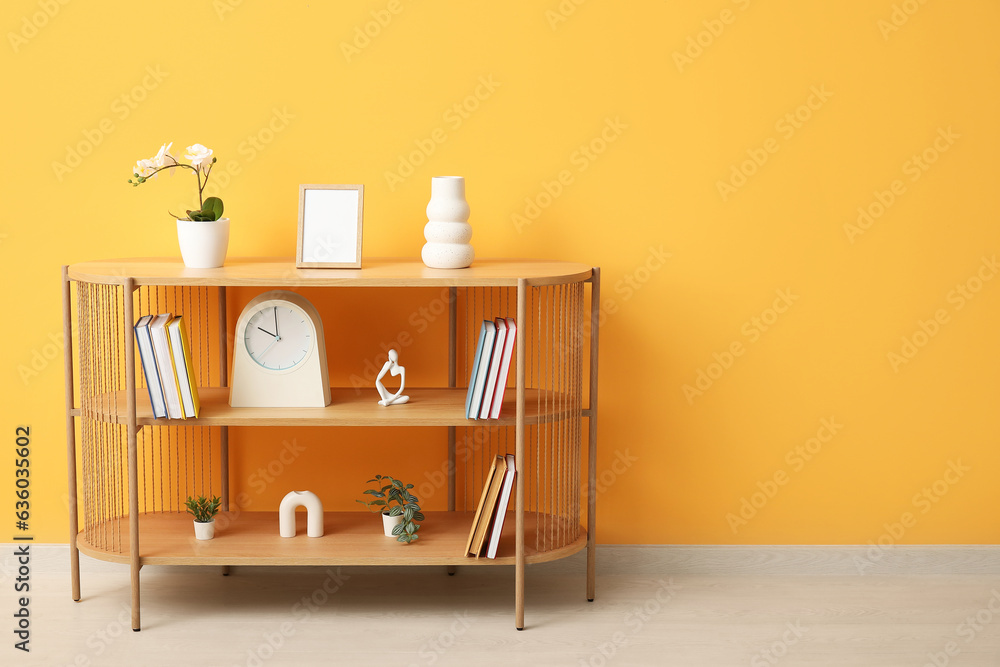 Shelving unit with books, orchid flower and decor near orange wall in room