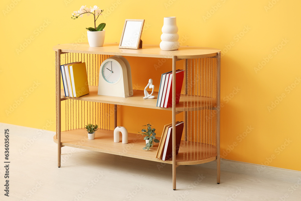 Shelving unit with books, orchid flower and decor near orange wall in room