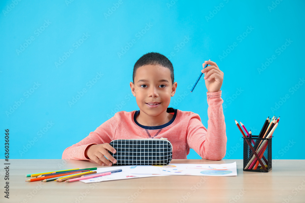 Little African-American boy with pencil case at table on blue background